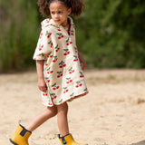 Child wearing the Little Green Radicals little cherries cream organic cotton kids hooded towel poncho.The poncho has a popper closure on the neck and a kangaroo pocket on the front. 