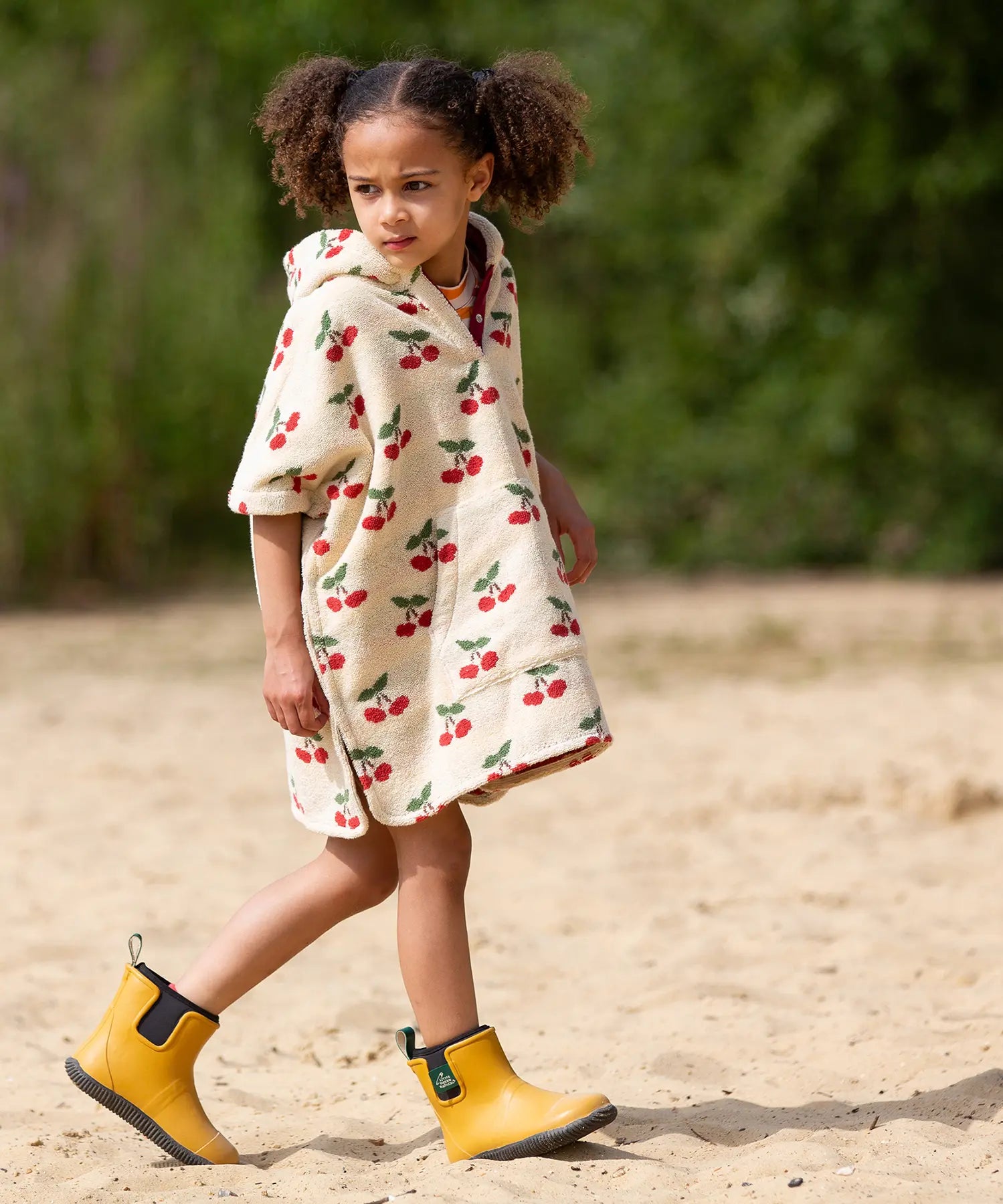 Child wearing the Little Green Radicals little cherries cream organic cotton kids hooded towel poncho.The poncho has a popper closure on the neck and a kangaroo pocket on the front. 
