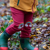 A close up of a child wearing the Little Green Radicals Red Rainbow Knee Patch Joggers in a forest. The close up shows the rainbow patches on the knees. Little green Radicals have a wide range of organic cotton, Fairtrade children's clothing an outerwear available here at Babipur.