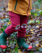 A close up of a child wearing the Little Green Radicals Red Rainbow Knee Patch Joggers in a forest. The close up shows the rainbow patches on the knees. Little green Radicals have a wide range of organic cotton, Fairtrade children's clothing an outerwear available here at Babipur.