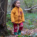 A child wearing the Little Green Radicals Red Rainbow Knee Patch Joggers with a yellow gold Polar Fleece Jacket and green wellingtons walking through a forest. Little green Radicals have a wide range of organic cotton, Fairtrade children's clothing an outerwear available here at Babipur
