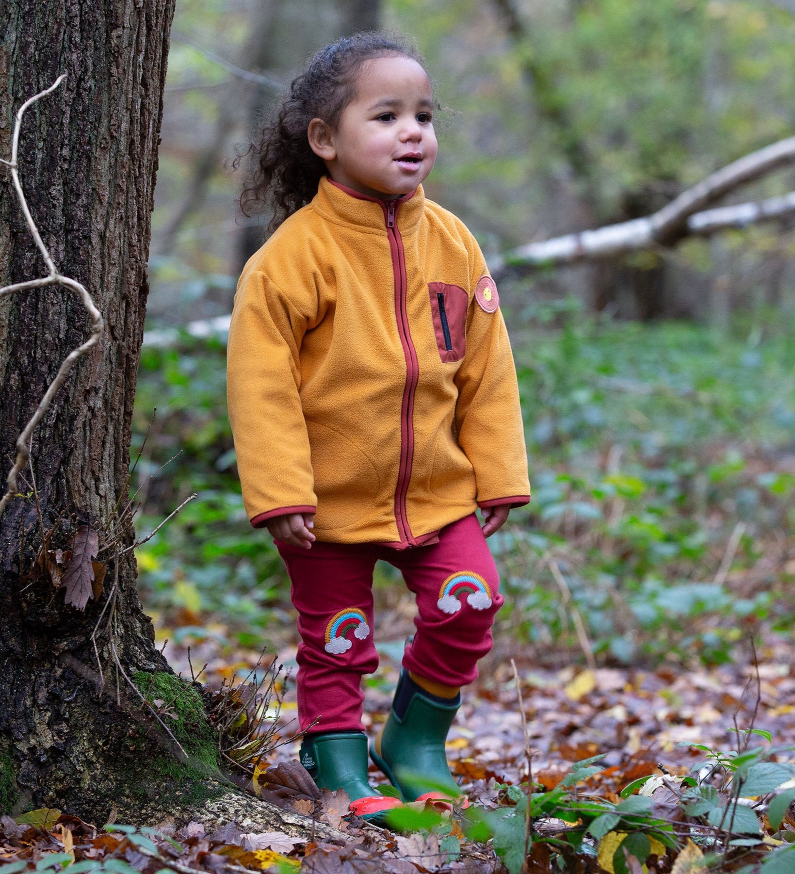 A child wearing the Little Green Radicals Red Rainbow Knee Patch Joggers with a yellow gold Polar Fleece Jacket and green wellingtons walking through a forest. Little green Radicals have a wide range of organic cotton, Fairtrade children's clothing an outerwear available here at Babipur