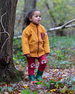 A child wearing the Little Green Radicals Red Rainbow Knee Patch Joggers with a yellow gold Polar Fleece Jacket and green wellingtons walking through a forest. Little green Radicals have a wide range of organic cotton, Fairtrade children's clothing an outerwear available here at Babipur