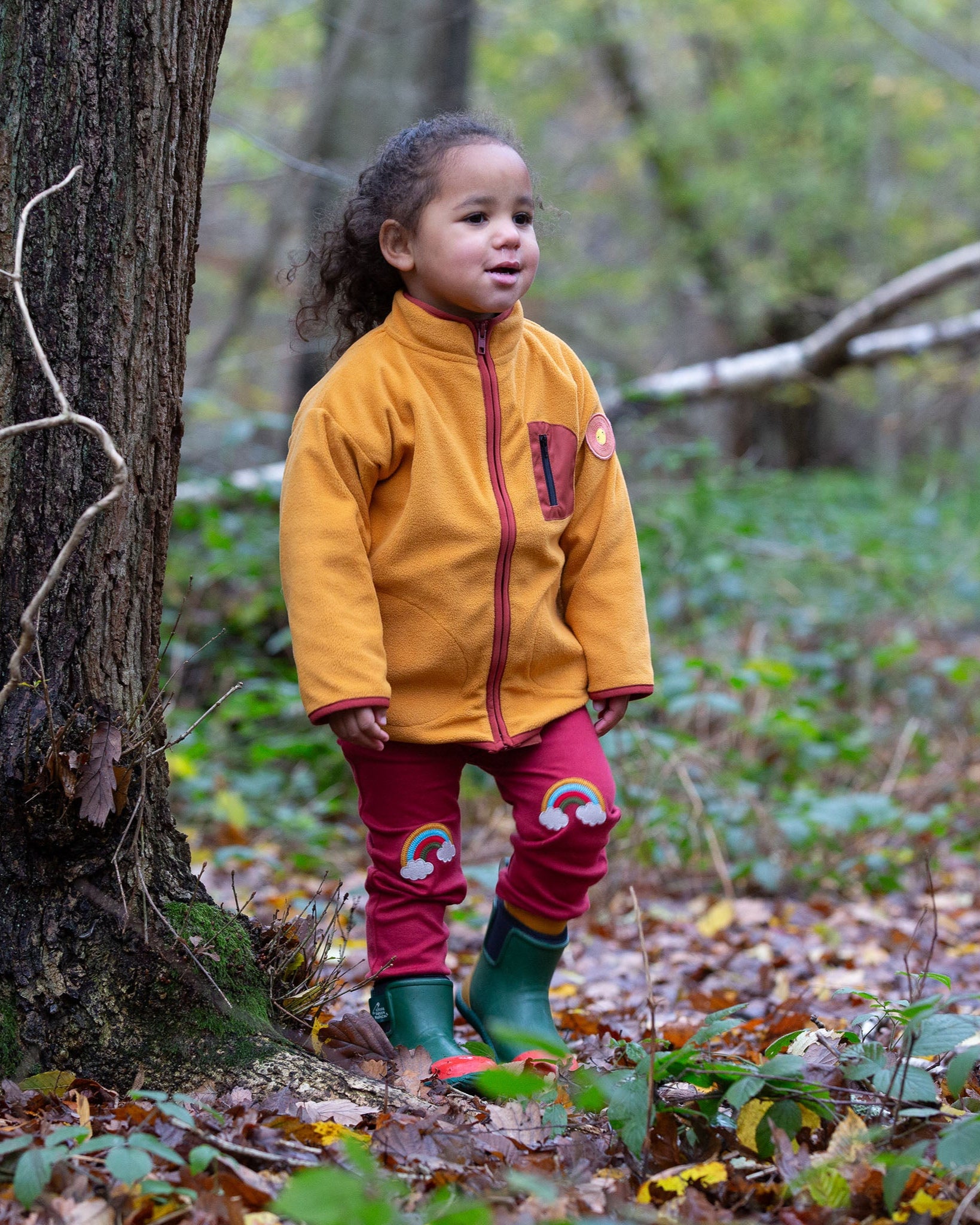 A child wearing the Little Green Radicals Red Rainbow Knee Patch Joggers with a yellow gold Polar Fleece Jacket and green wellingtons walking through a forest. Little green Radicals have a wide range of organic cotton, Fairtrade children's clothing an outerwear available here at Babipur