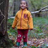 A child wearing the Little Green Radicals Red Rainbow Knee Patch Joggers with a yellow gold Polar Fleece Jacket and green wellingtons walking through a forest. Little green Radicals have a wide range of organic cotton, Fairtrade children's clothing an outerwear available here at Babipur
