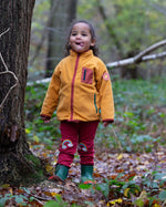 A child wearing the Little Green Radicals Red Rainbow Knee Patch Joggers with a yellow gold Polar Fleece Jacket and green wellingtons walking through a forest. Little green Radicals have a wide range of organic cotton, Fairtrade children's clothing an outerwear available here at Babipur