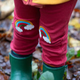 A close up of a child wearing the Little Green Radicals Red Rainbow Knee Patch Joggers in a forest. The close up shows the rainbow patches on the knees. Little green Radicals have a wide range of organic cotton, Fairtrade children's clothing an outerwear available here at Babipur.