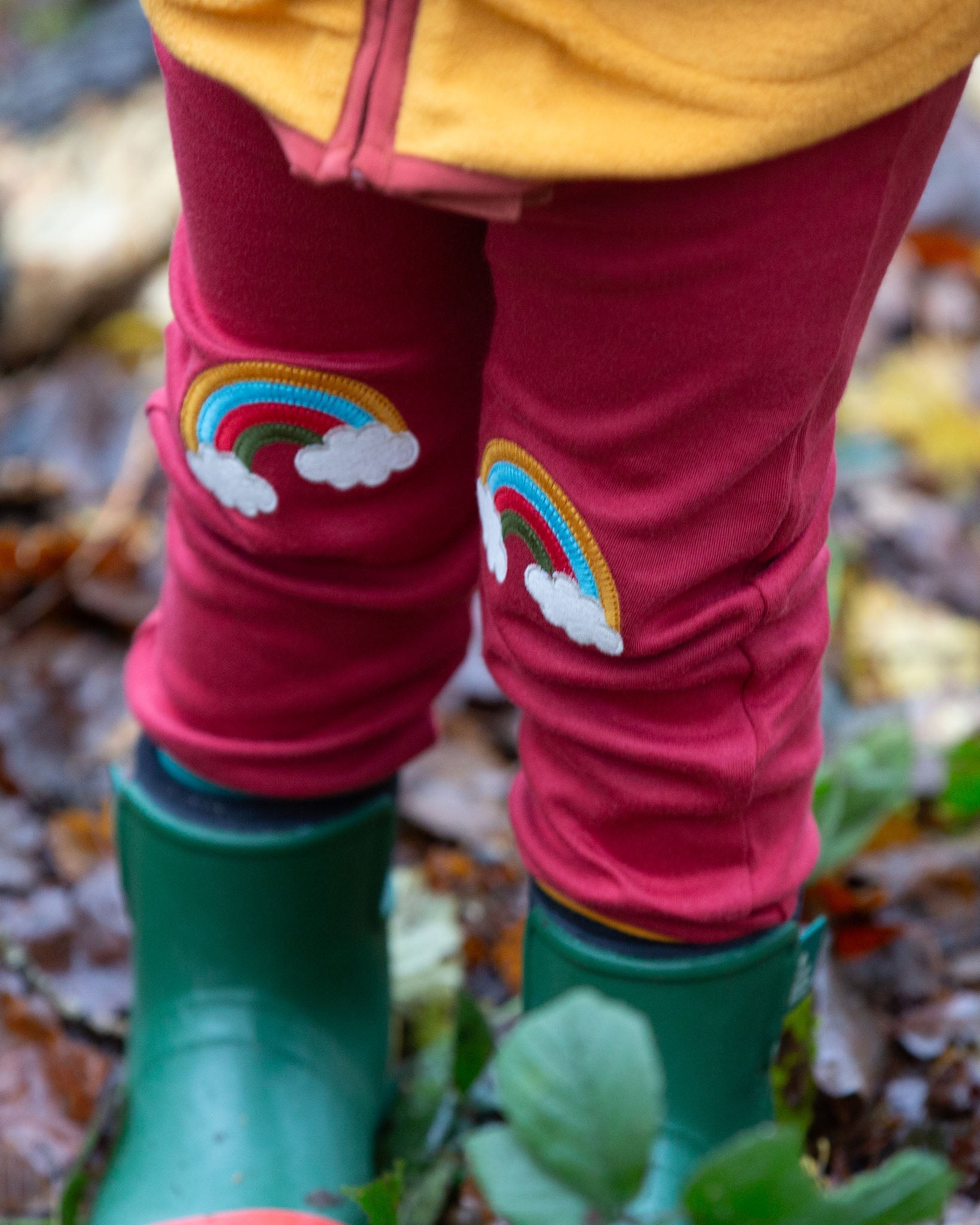 A close up of a child wearing the Little Green Radicals Red Rainbow Knee Patch Joggers in a forest. The close up shows the rainbow patches on the knees. Little green Radicals have a wide range of organic cotton, Fairtrade children's clothing an outerwear available here at Babipur.