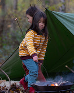 A child standing by a campfire in the forest, wearing the Little Green Radicals Reversible Easy Rider Jacket on the Brown Bears print side. Little green Radicals have a wide range of organic cotton, Fairtrade children's clothing an outerwear available here at Babipur.