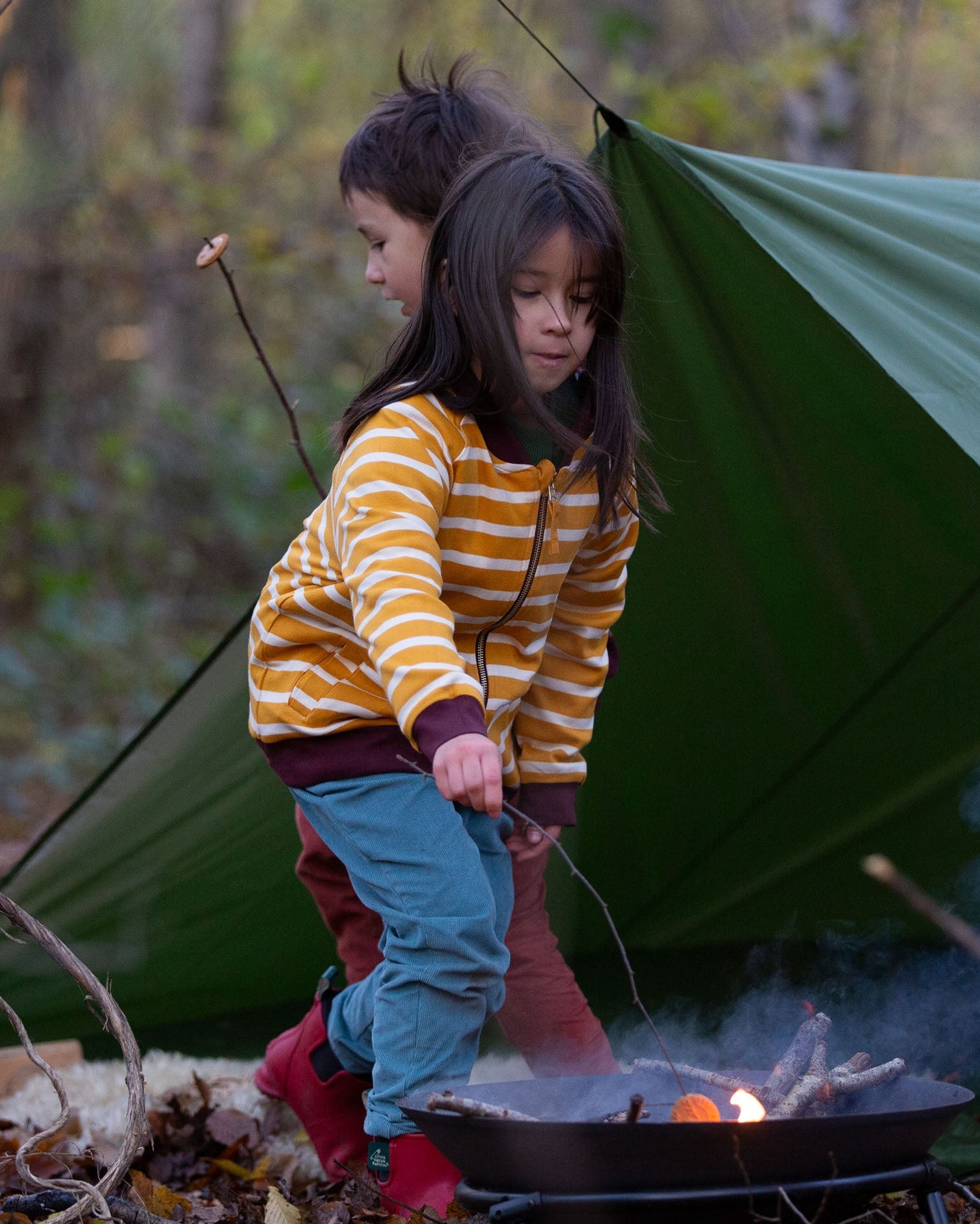 A child standing by a campfire in the forest, wearing the Little Green Radicals Reversible Easy Rider Jacket on the Brown Bears print side. Little green Radicals have a wide range of organic cotton, Fairtrade children's clothing an outerwear available here at Babipur.