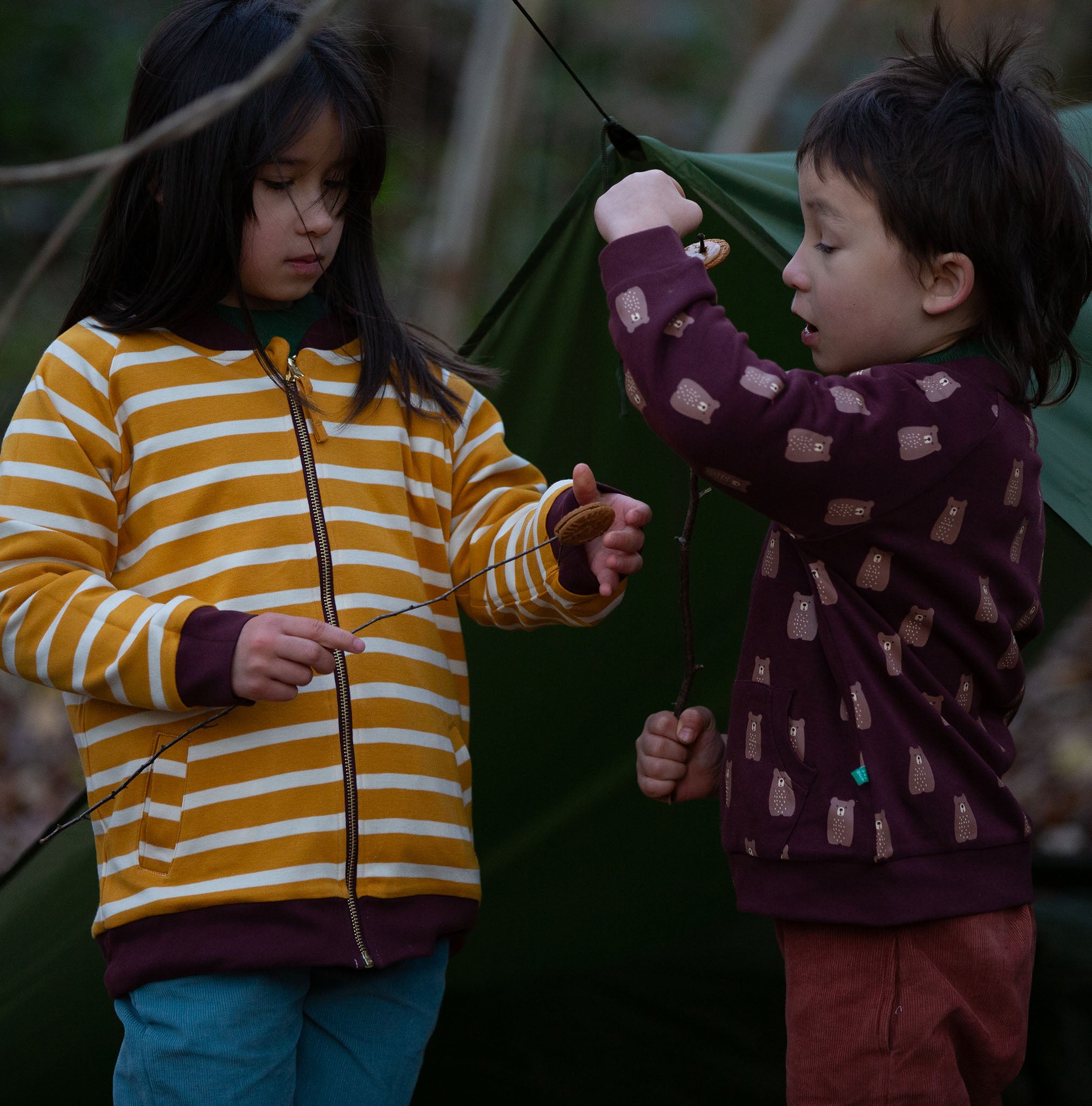 Two children standing in front of a green tent in the forest. Both are wearing the Little Green Radicals Reversible Easy Rider Jacket. One on the Brown Bears print side the other on a yellow gold and white stripe side. Little green Radicals have a wide range of organic cotton, Fairtrade children's clothing an outerwear available here at Babipur