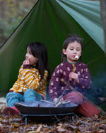 Two children sitting besides a campfire in the forest. Both are wearing the Little Green Radicals Reversible Easy Rider Jacket. One on the Brown Bears print side the other on a yellow gold and white stripe side. Little green Radicals have a wide range of organic cotton, Fairtrade children's clothing an outerwear available here at Babipur
