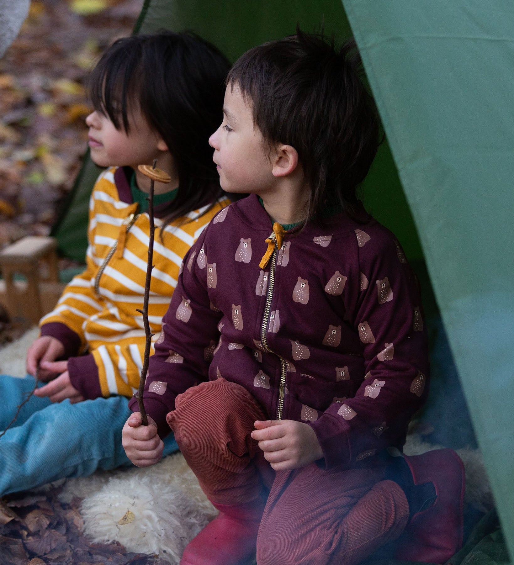 Two children sitting in a tent in the forest. Both are wearing the Little Green Radicals Reversible Easy Rider Jacket. One on the Brown Bears print side the other on a yellow gold and white stripe side. Little green Radicals have a wide range of organic cotton, Fairtrade children's clothing an outerwear available here at Babipur