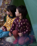 Two children sitting in a tent in the forest. Both are wearing the Little Green Radicals Reversible Easy Rider Jacket. One on the Brown Bears print side the other on a yellow gold and white stripe side. Little green Radicals have a wide range of organic cotton, Fairtrade children's clothing an outerwear available here at Babipur