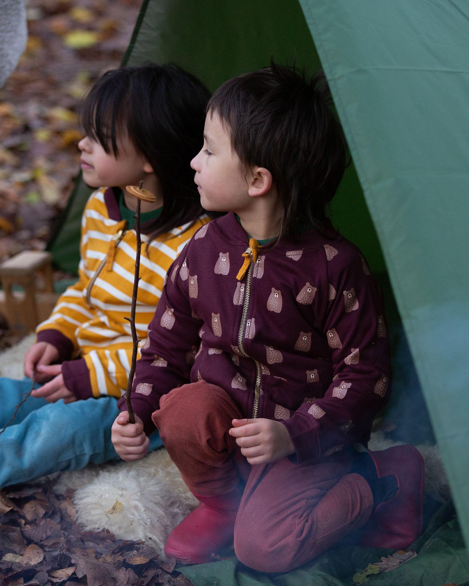 Two children sitting in a tent in the forest. Both are wearing the Little Green Radicals Reversible Easy Rider Jacket. One on the Brown Bears print side the other on a yellow gold and white stripe side. Little green Radicals have a wide range of organic cotton, Fairtrade children's clothing an outerwear available here at Babipur