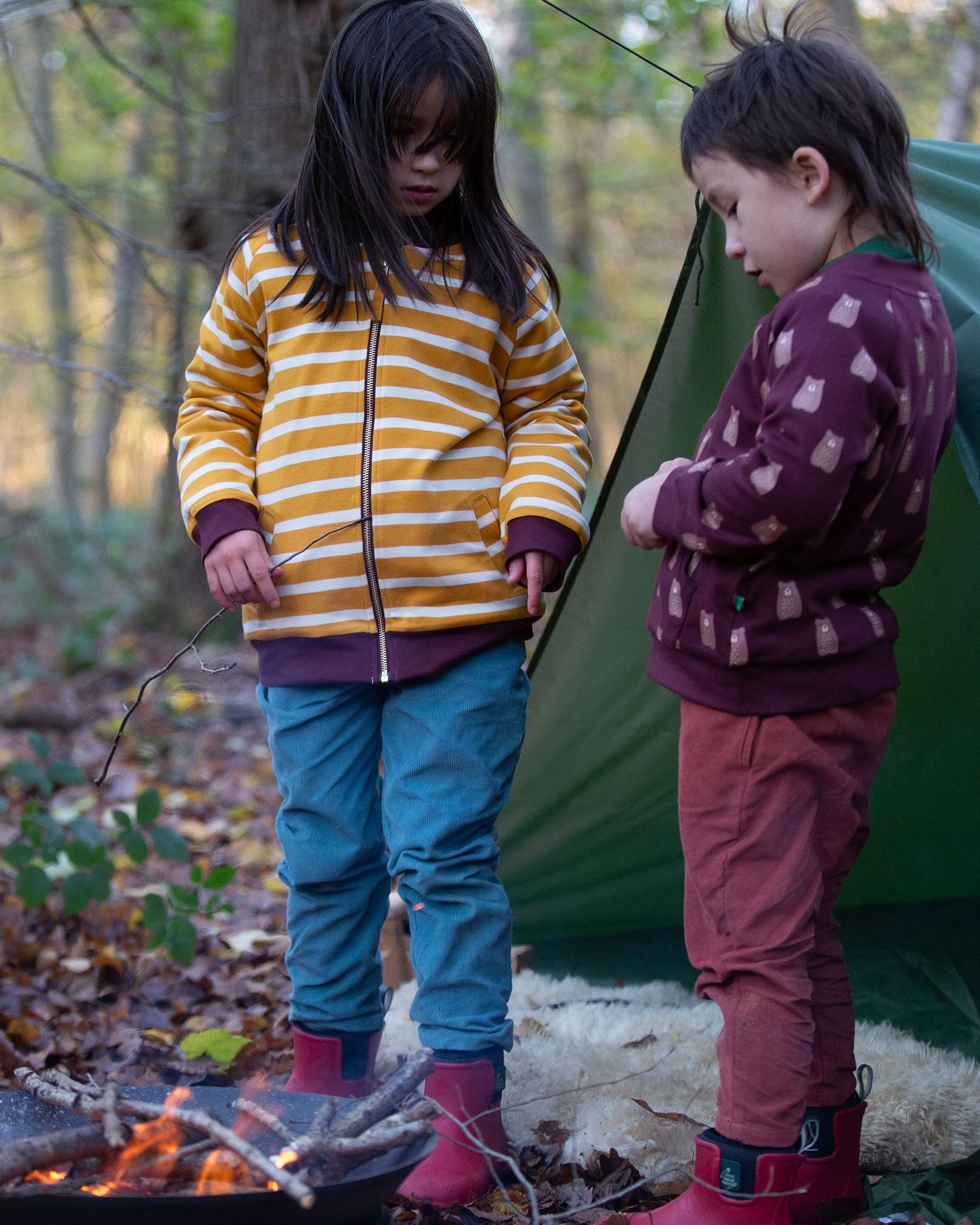 Two children standing in front of a tent in the forest. Both are wearing the Little Green Radicals Reversible Easy Rider Jacket. One on the Brown Bears print side the other on a yellow gold and white stripe side. Little green Radicals have a wide range of organic cotton, Fairtrade children's clothing an outerwear available here at Babipur.