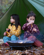 Two children sitting besides a campfire in the forest. Both are wearing the Little Green Radicals Reversible Easy Rider Jacket. One on the Brown Bears print side the other on a yellow gold and white stripe side. Little green Radicals have a wide range of organic cotton, Fairtrade children's clothing an outerwear available here at Babipur