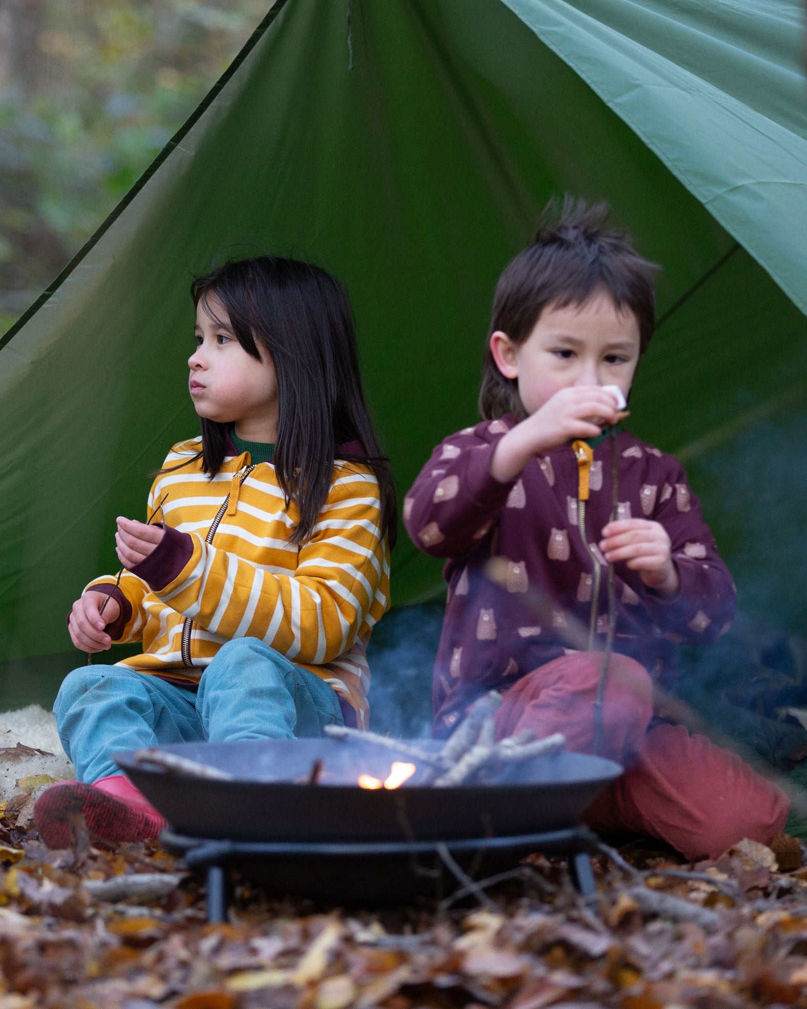 Two children sitting besides a campfire in the forest. Both are wearing the Little Green Radicals Reversible Easy Rider Jacket. One on the Brown Bears print side the other on a yellow gold and white stripe side. Little green Radicals have a wide range of organic cotton, Fairtrade children's clothing an outerwear available here at Babipur
