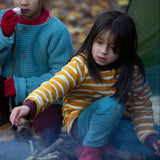 A child kneeling down near a campfire in the forest wearing the Little Green Radicals Reversible Easy Rider Jacket on the  yellow gold and white stripe side. Little green Radicals have a wide range of organic cotton, Fairtrade children's clothing an outerwear available here at Babipur