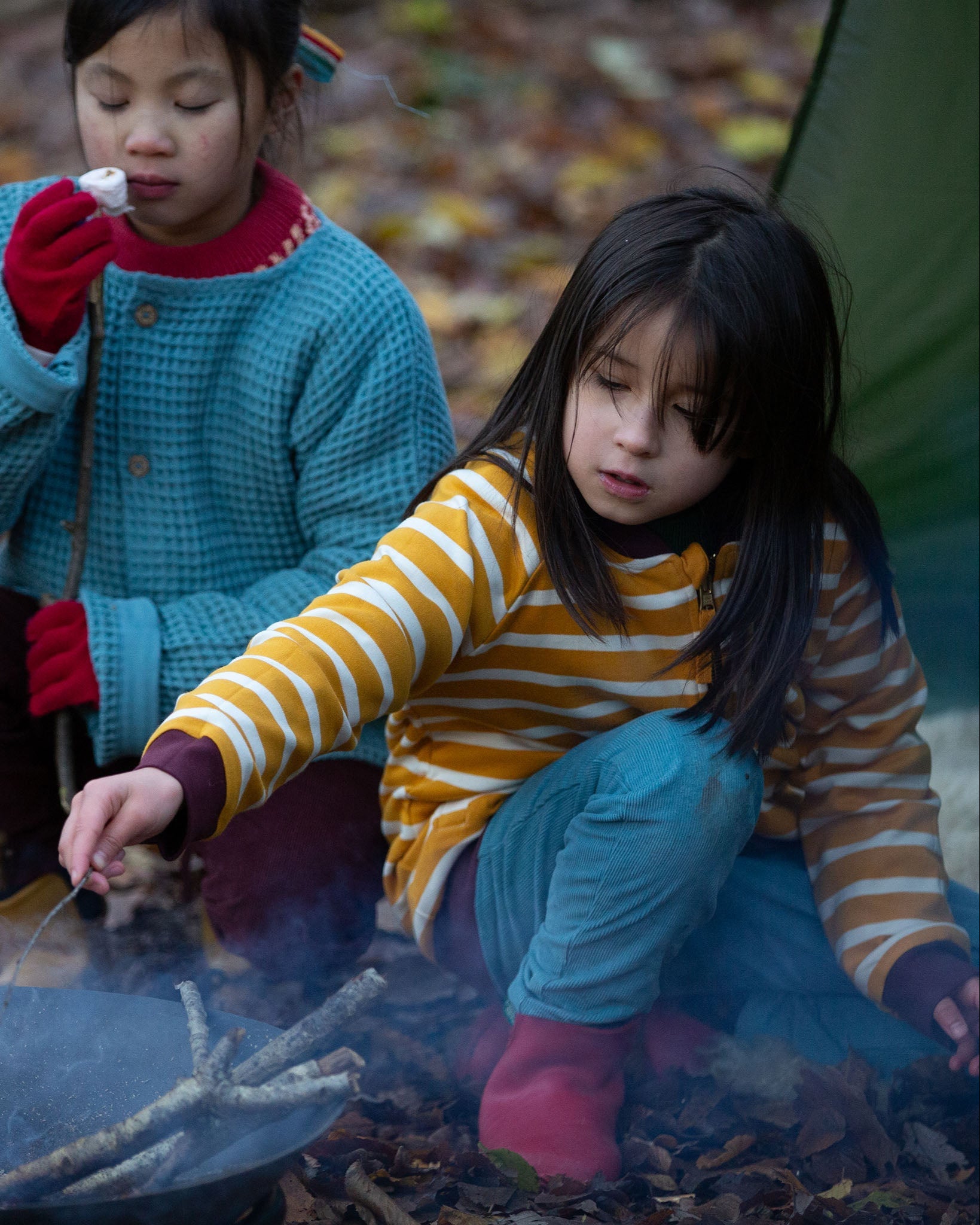 A child kneeling down near a campfire in the forest wearing the Little Green Radicals Reversible Easy Rider Jacket on the  yellow gold and white stripe side. Little green Radicals have a wide range of organic cotton, Fairtrade children's clothing an outerwear available here at Babipur
