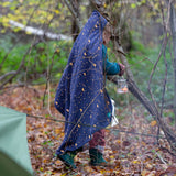 A child walking through a forest wearing a Little Green Radicals dark night print Organic Sherpa Hooded Baby Blanket over their head.  A GOTS organic cotton navy blanket with a starry night print and a brand logo patch on the corner. Features a white sherpa style fleece lining. This blanket is a part of a wide range of organic cotton clothing and accessories for children available here at Babipur. 