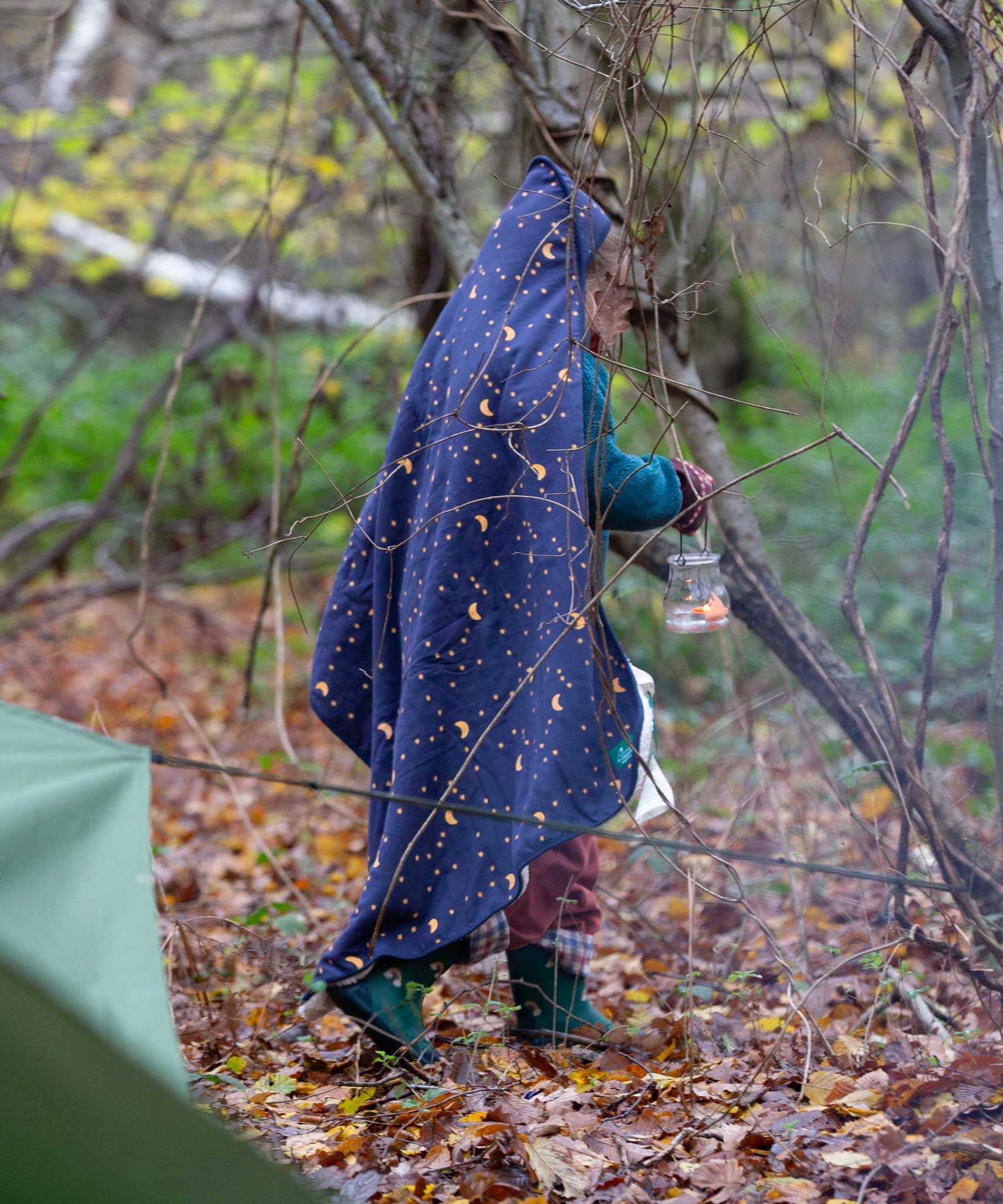 A child walking through a forest wearing a Little Green Radicals dark night print Organic Sherpa Hooded Baby Blanket over their head.  A GOTS organic cotton navy blanket with a starry night print and a brand logo patch on the corner. Features a white sherpa style fleece lining. This blanket is a part of a wide range of organic cotton clothing and accessories for children available here at Babipur. 
