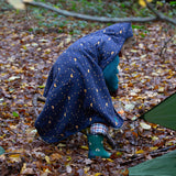 A child walking through a forest wearing a Little Green Radicals dark night print Organic Sherpa Hooded Baby Blanket over their head.  A GOTS organic cotton navy blanket with a starry night print and a brand logo patch on the corner. Features a white sherpa style fleece lining. This blanket is a part of a wide range of organic cotton clothing and accessories for children available here at Babipur. 