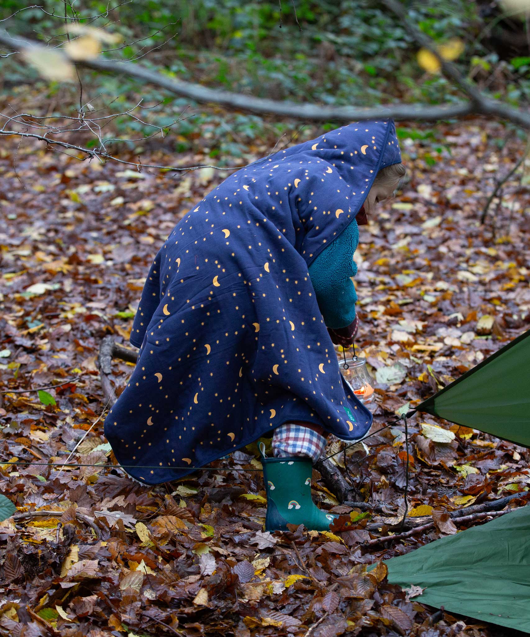 A child walking through a forest wearing a Little Green Radicals dark night print Organic Sherpa Hooded Baby Blanket over their head.  A GOTS organic cotton navy blanket with a starry night print and a brand logo patch on the corner. Features a white sherpa style fleece lining. This blanket is a part of a wide range of organic cotton clothing and accessories for children available here at Babipur. 
