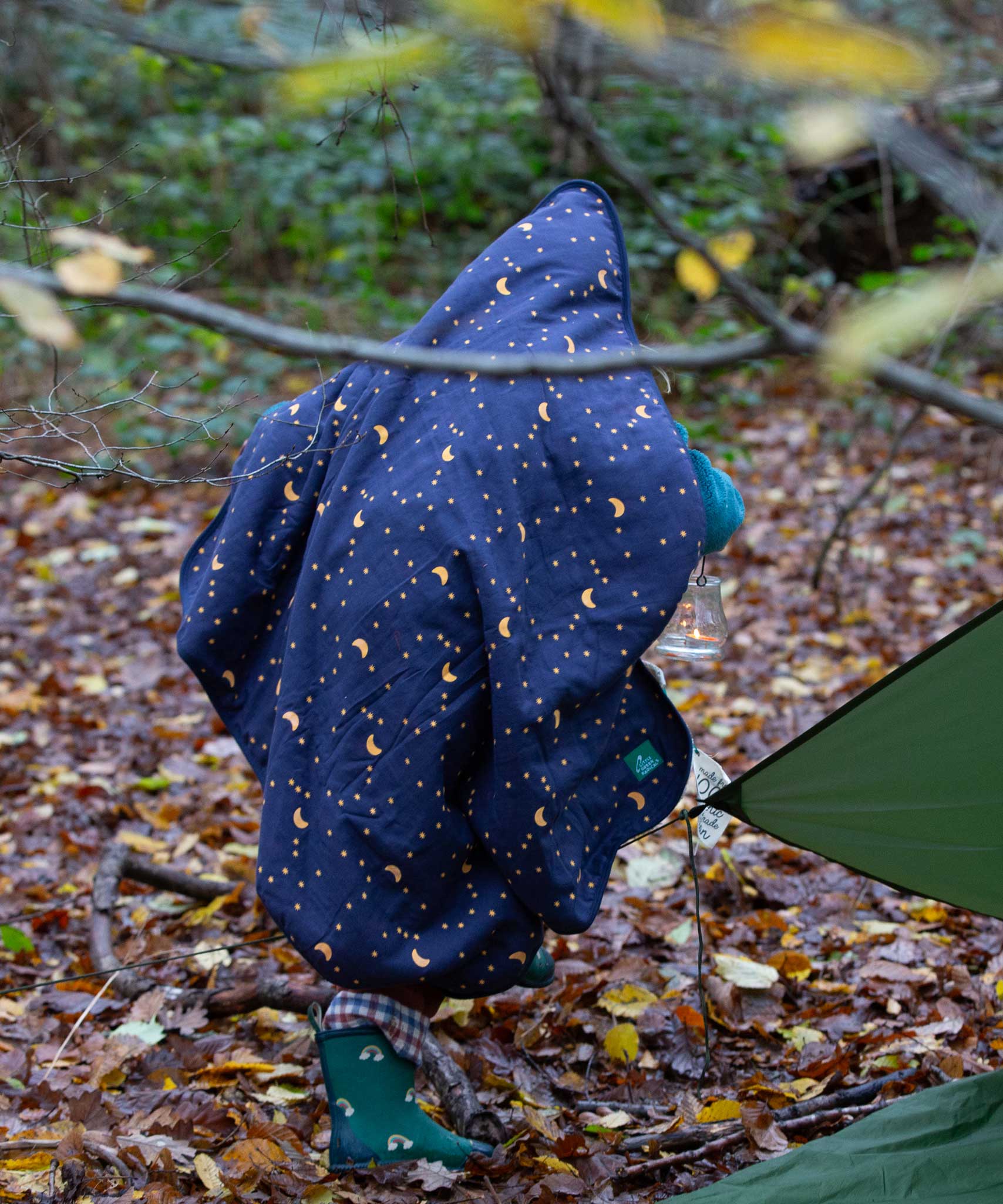 A child walking through a forest wearing a Little Green Radicals dark night print Organic Sherpa Hooded Baby Blanket over their head.  A GOTS organic cotton navy blanket with a starry night print and a brand logo patch on the corner. Features a white sherpa style fleece lining. This blanket is a part of a wide range of organic cotton clothing and accessories for children available here at Babipur. 
