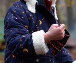 A close up of a child's hands wearing the Little Green Radicals Kids Dark Night design Organic Sherpa lined Toasty Coat. This warm coat is navy with a yellow moon and stars print all over with a white sherpa fleece lining. This coat is a part of a wide range of organic cotton clothing for children available here at Babipur. 