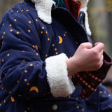 A close up of a child's hands wearing the Little Green Radicals Kids Dark Night design Organic Sherpa lined Toasty Coat. This warm coat is navy with a yellow moon and stars print all over with a white sherpa fleece lining. This coat is a part of a wide range of organic cotton clothing for children available here at Babipur. 