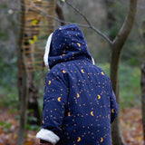 A view of the back of a child wearing the Little Green Radicals Kids Dark Night design Organic Sherpa lined Toasty Coat. The child has the hood up. This warm coat is navy with a yellow moon and stars print all over with a white sherpa fleece lining. This coat is a part of a wide range of organic cotton clothing for children available here at Babipur. 