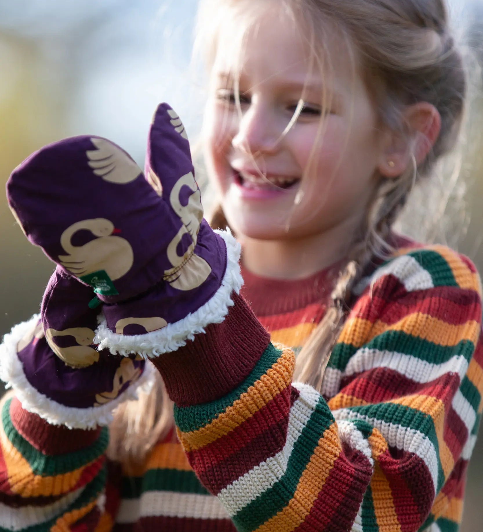 Child wearing Little Green Radicals  sleeping swans print sherpa fleece lined purple mittens with a rainbow striped knitted jumper.
