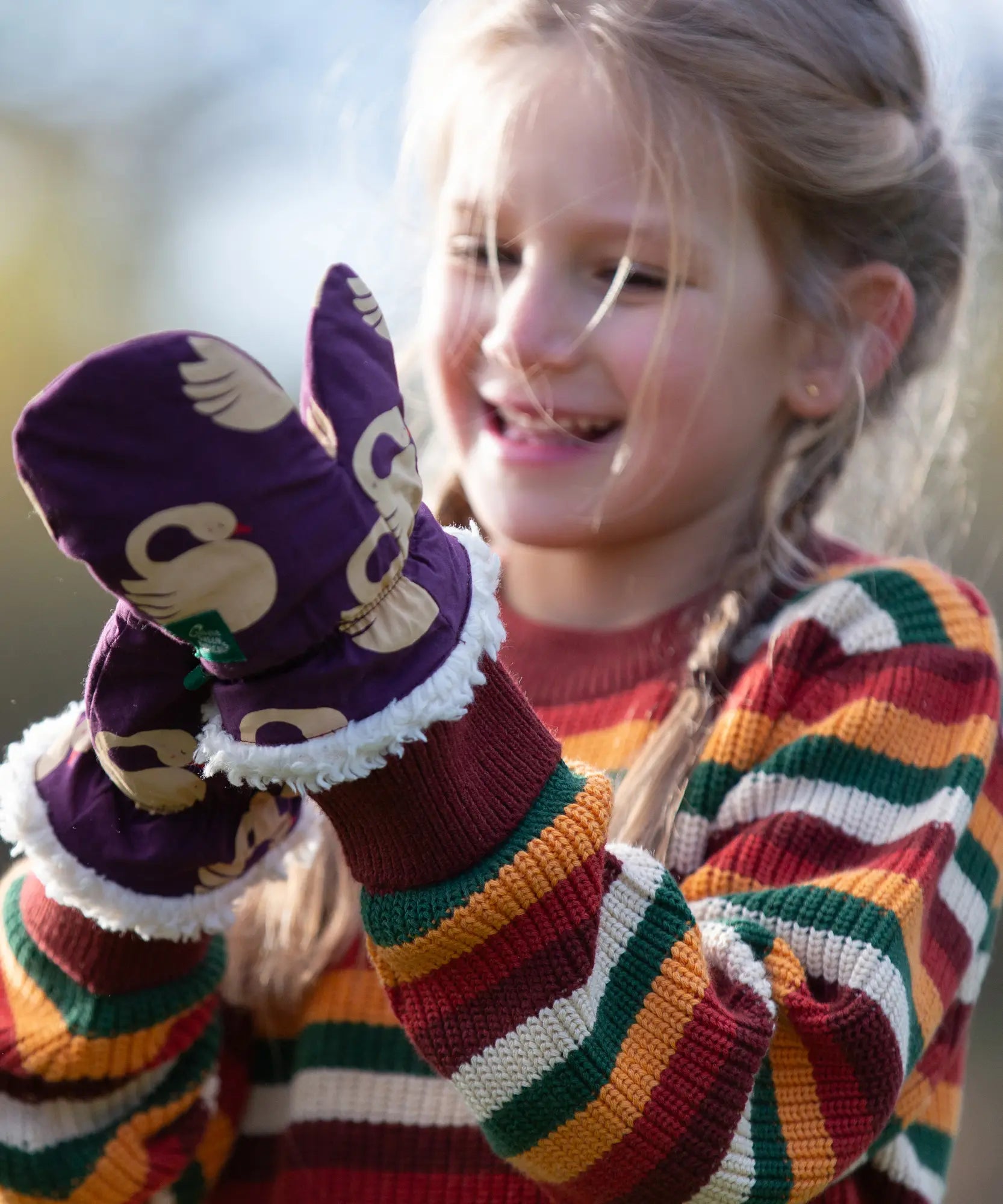 Child wearing Little Green Radicals  sleeping swans print sherpa fleece lined purple mittens with a rainbow striped knitted jumper.