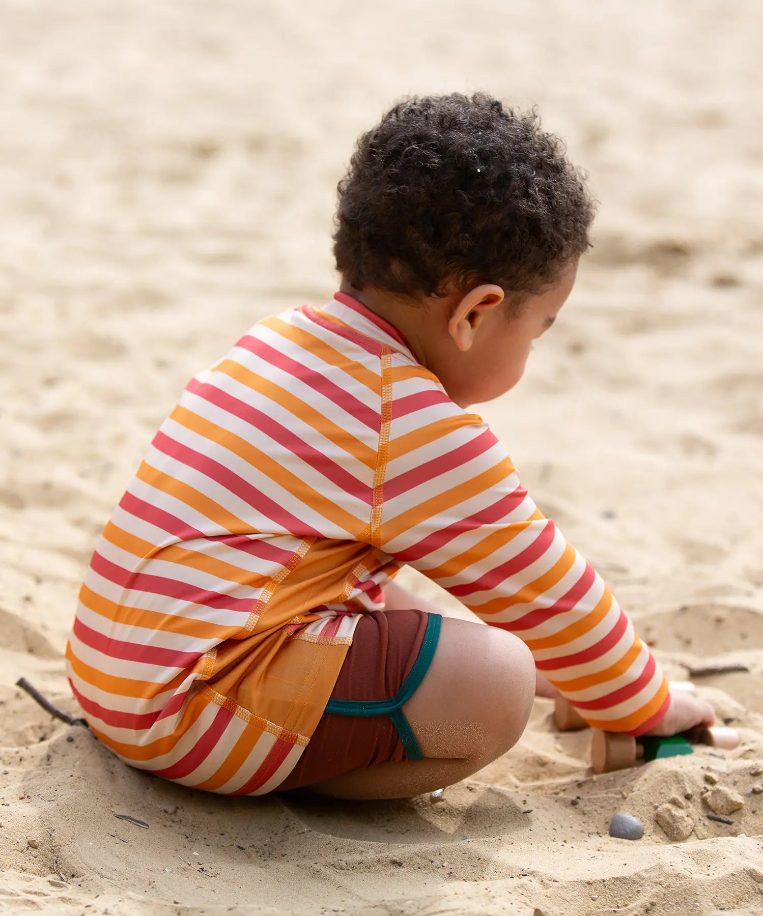 Back of a child wearing the Little Green Radicals sorbet striped UPF 50 sunsafe long sleeve rash vest with dark red swim shorts.