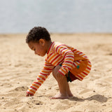 Child on beach wearing the Little Green Radicals sorbet striped UPF 50 sunsafe long sleeve rash vest with dark red swim shorts.  