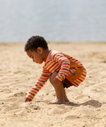 Child on beach wearing the Little Green Radicals sorbet striped UPF 50 sunsafe long sleeve rash vest with dark red swim shorts.  