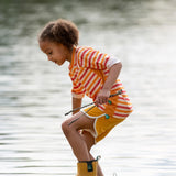 Child wearing the Little Green Radicals sorbet striped UPF 50 sunsafe long sleeve rash vest with yellow swim shorts and wellington boots walking through shallow water.