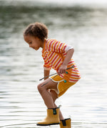 Child wearing the Little Green Radicals sorbet striped UPF 50 sunsafe long sleeve rash vest with yellow swim shorts and wellington boots walking through shallow water.