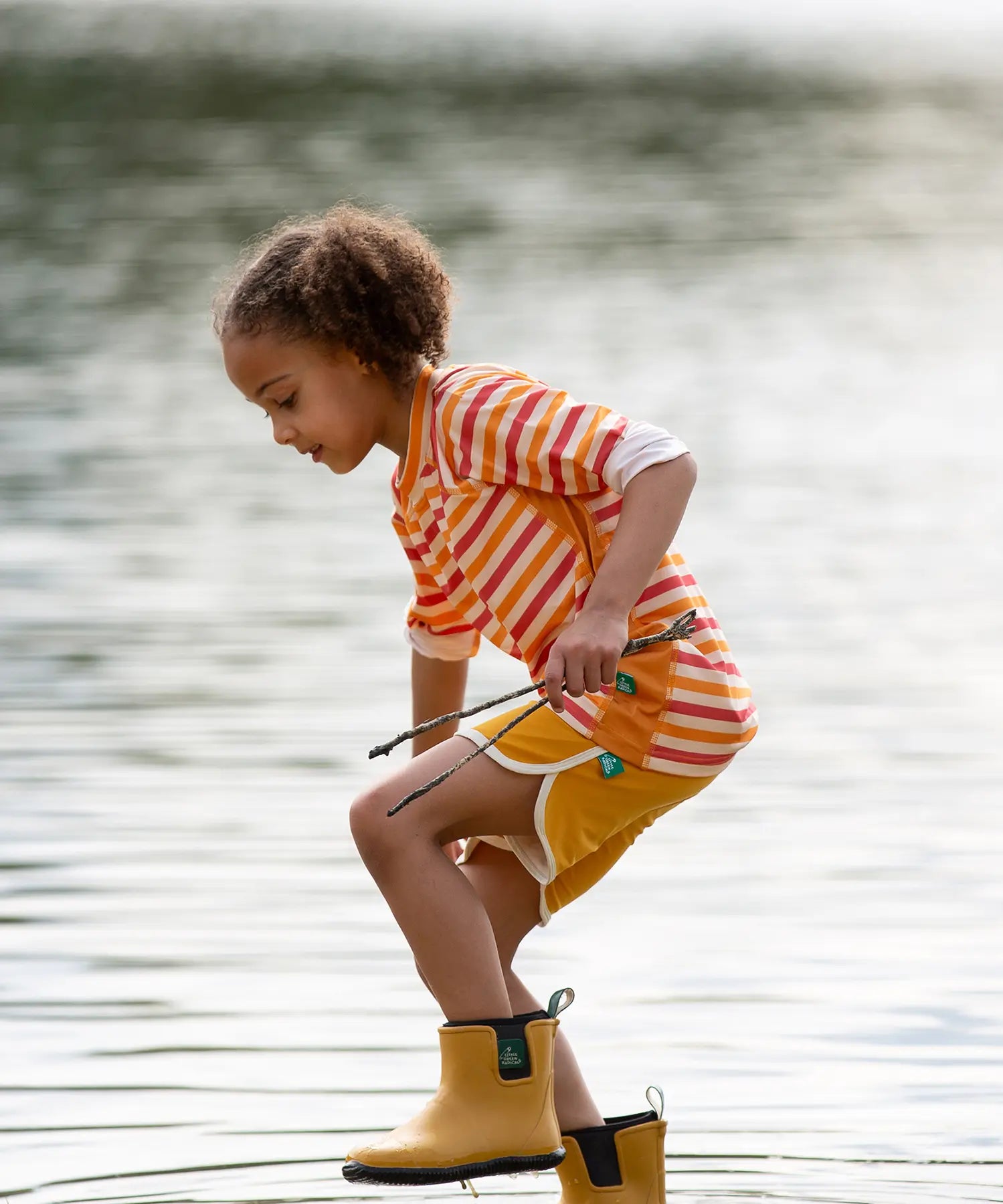 Child wearing the Little Green Radicals sorbet striped UPF 50 sunsafe long sleeve rash vest with yellow swim shorts and wellington boots walking through shallow water.