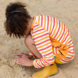 Child kneeling on beach wearing the Little Green Radicals sorbet striped UPF 50 sunsafe long sleeve rash vest  with yellow gold swim shorts and yellow gold wellington boots.