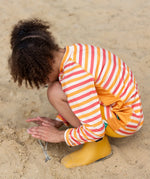 Child kneeling on beach wearing the Little Green Radicals sorbet striped UPF 50 sunsafe long sleeve rash vest  with yellow gold swim shorts and yellow gold wellington boots.