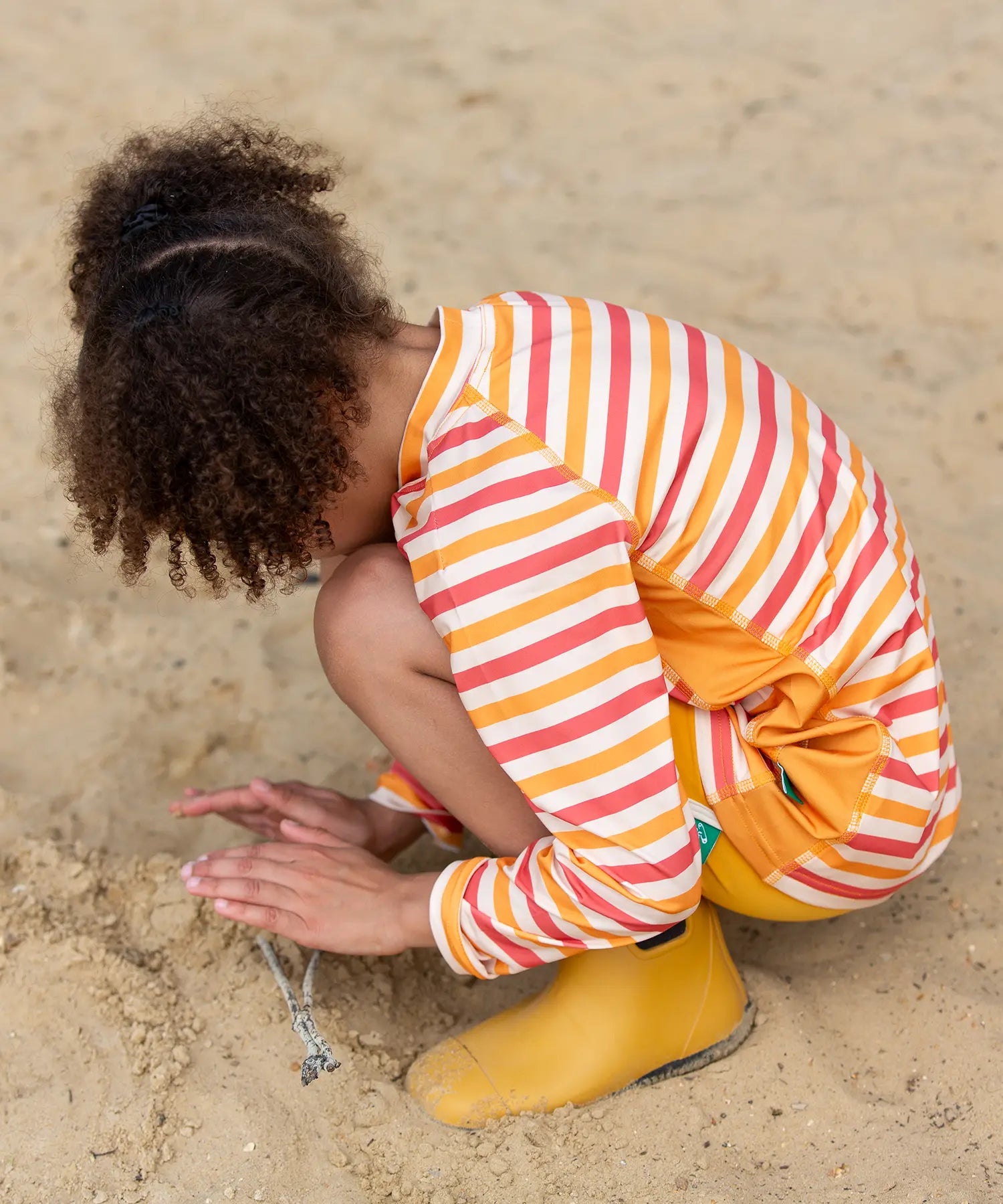 Child kneeling on beach wearing the Little Green Radicals sorbet striped UPF 50 sunsafe long sleeve rash vest  with yellow gold swim shorts and yellow gold wellington boots.
