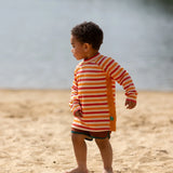 Child on beach wearing the Little Green Radicals sorbet striped UPF 50 sunsafe long sleeve rash vest with dark red swim shorts.
