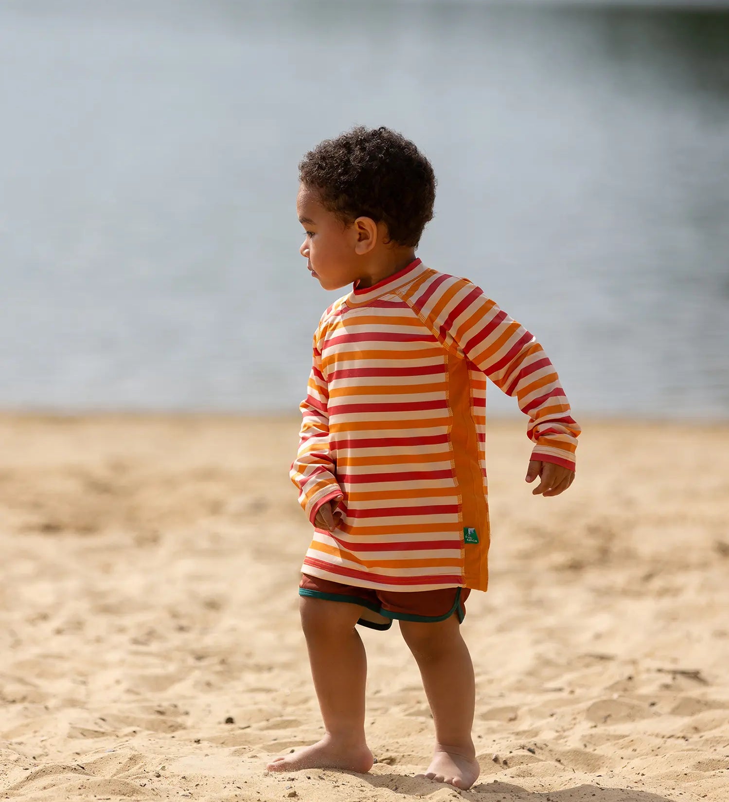 Child on beach wearing the Little Green Radicals sorbet striped UPF 50 sunsafe long sleeve rash vest with dark red swim shorts.