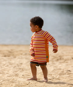 Child on beach wearing the Little Green Radicals sorbet striped UPF 50 sunsafe long sleeve rash vest with dark red swim shorts.