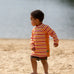 Child on beach wearing the Little Green Radicals sorbet striped UPF 50 sunsafe long sleeve rash vest with dark red swim shorts.