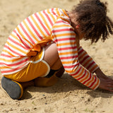 Child wearing the Little Green Radicals sorbet striped UPF 50 sunsafe long sleeve rash vest  with yellow swim shorts and wellington boots.