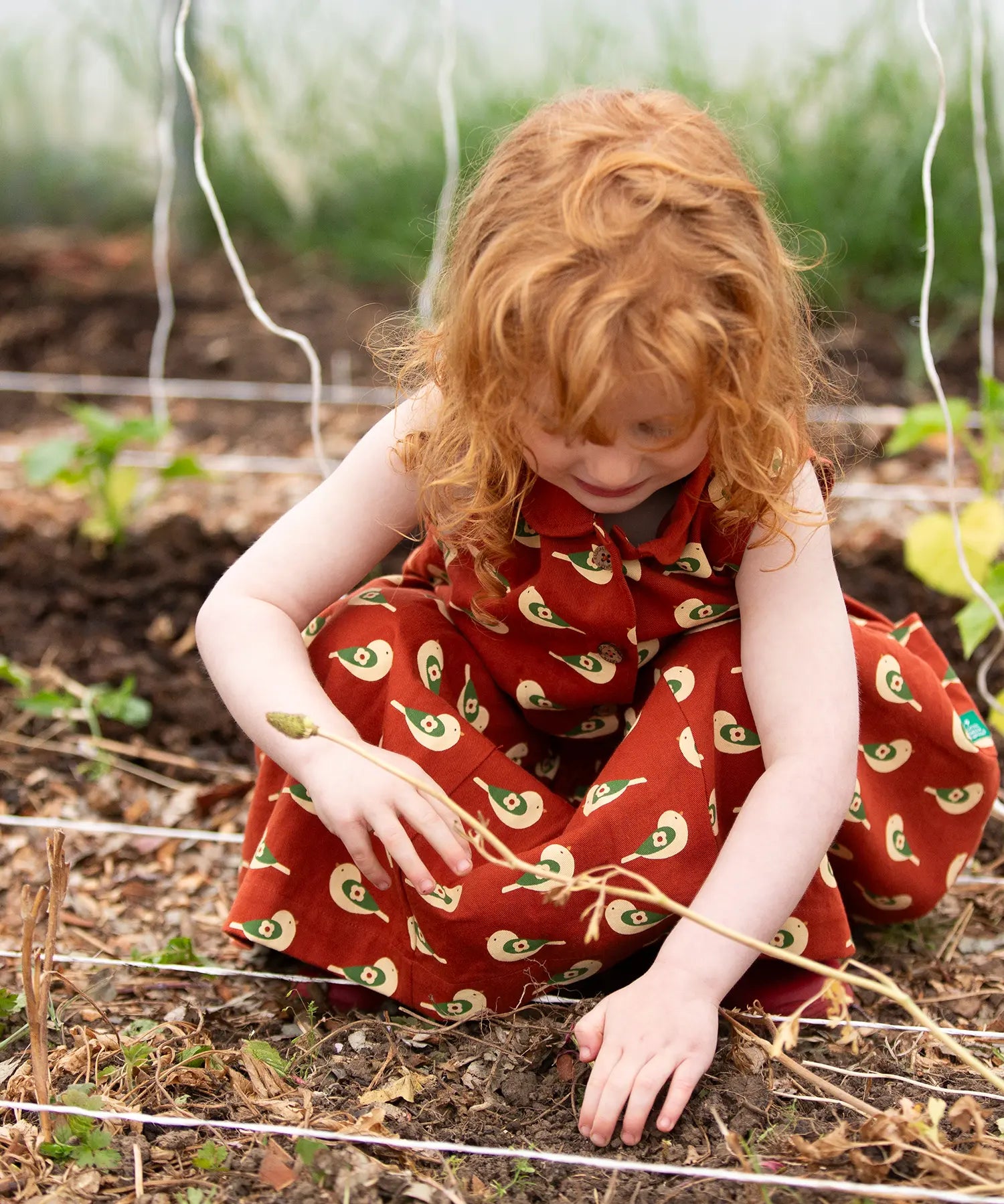 Child kneeling down wearing the Little Green Radicals child's red repeated spring birds print collared pinafore button dress. 
