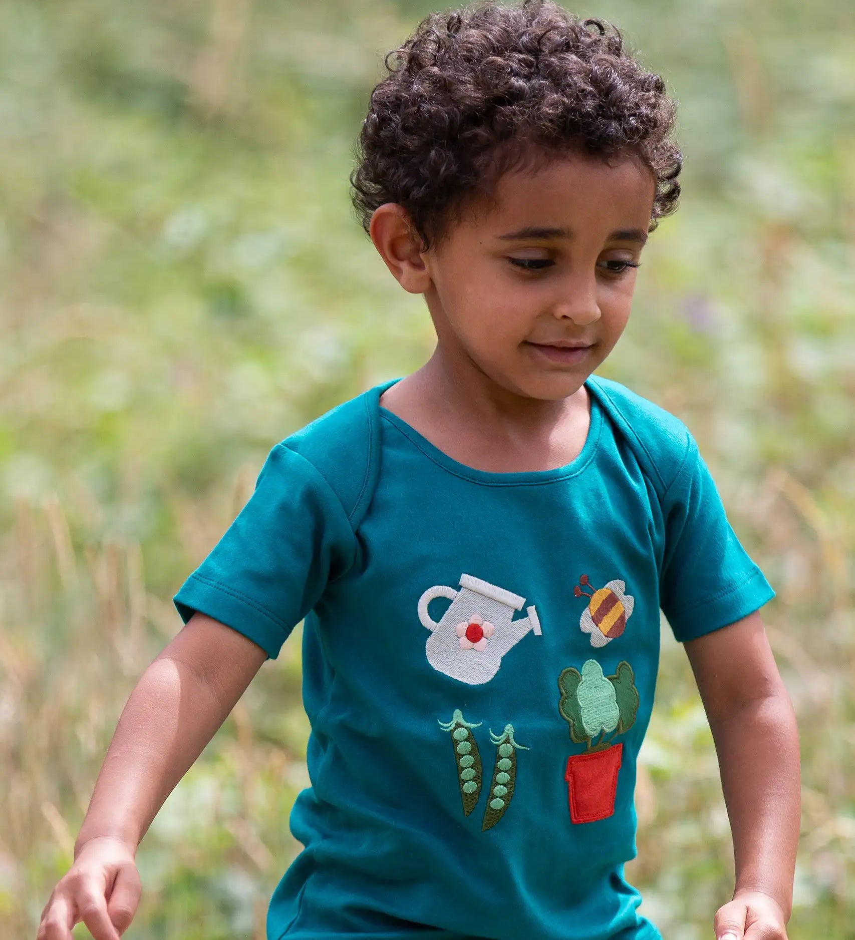 Child wearing the Little Green Radicals envelope neck Spring garden embroidered organic cotton green t-shirt. Features 4 embroidered elements, a white watering can, peas in their pods, a bee and a potted plant.